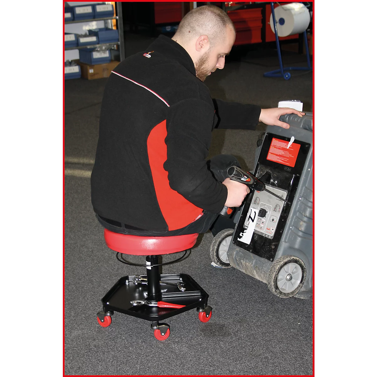 Un homme est assis sur un tabouret rouge à roulettes et travaille sur un appareil. Il porte des vêtements noirs.